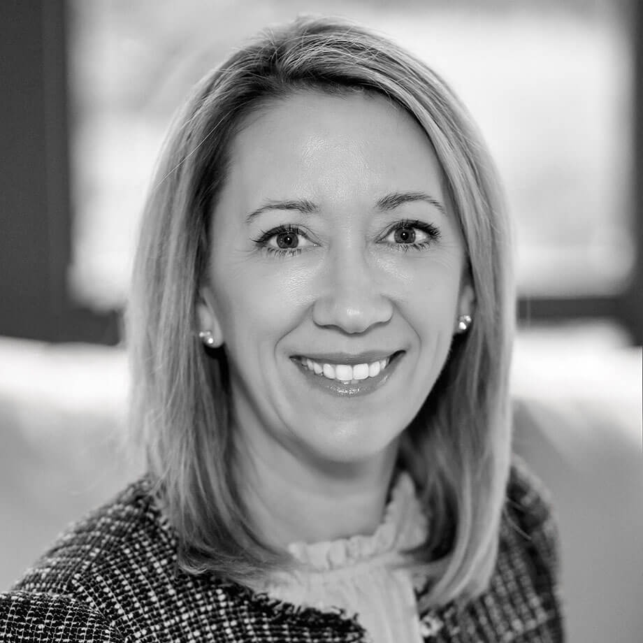 Professional headshot of Kate McBreen, smiling confidently at the camera, wearing a textured jacket, in black and white