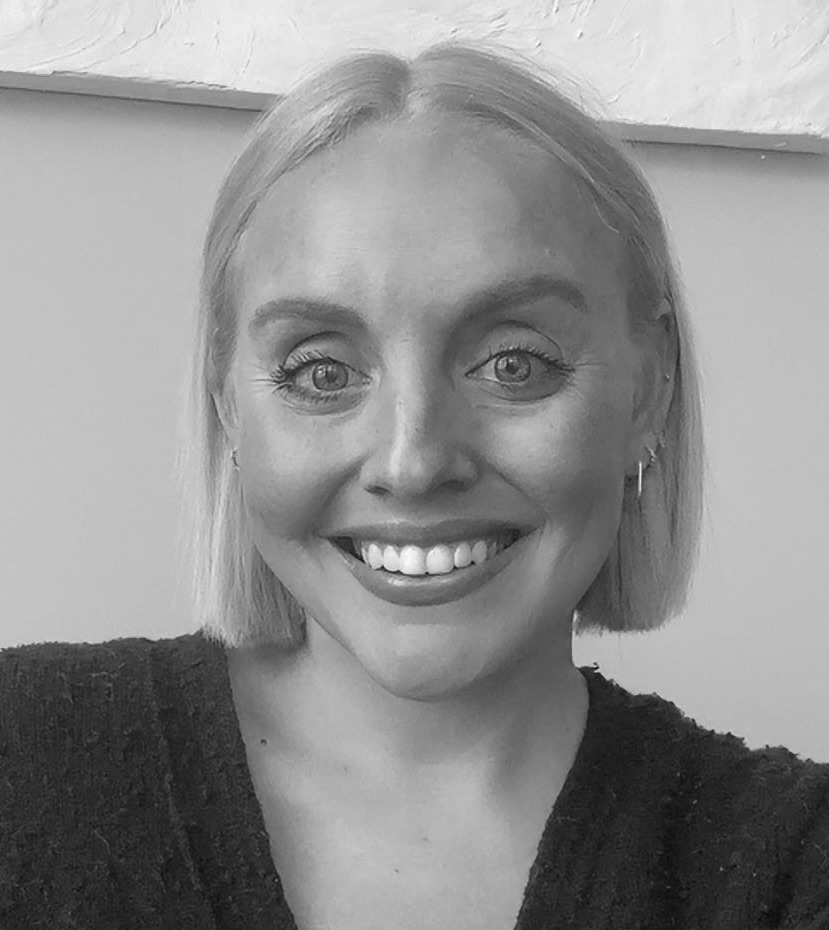Portrait of Alex Belden, a woman with straight chin-length blonde hair, smiling broadly at the camera, wearing small stud earrings and a dark textured cardigan against a plain light wall, in black and white